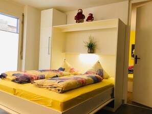 Een bed met een geel matras en kleurrijke kussens, een nachtkastje met een lamp, en een plank met twee teddyberen en een plant.