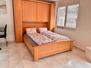 Ein Einzelbett mit Holzbettrahmen und einer blumigen Tagesdecke in einem Zimmer mit gefliestem Boden und einem Fenster mit Spitzenvorhängen.