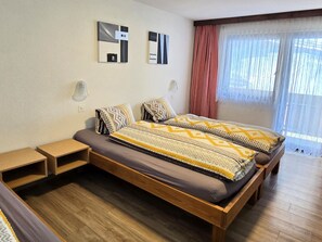 A modern bedroom with a wooden bed, two bedside tables, a nightstand, and a window with sheer curtains. - Ferienhaus Adonis Appartement/Fewo 5-Bettwohnung "Weissmies" by Interhome (Saas-Grund)