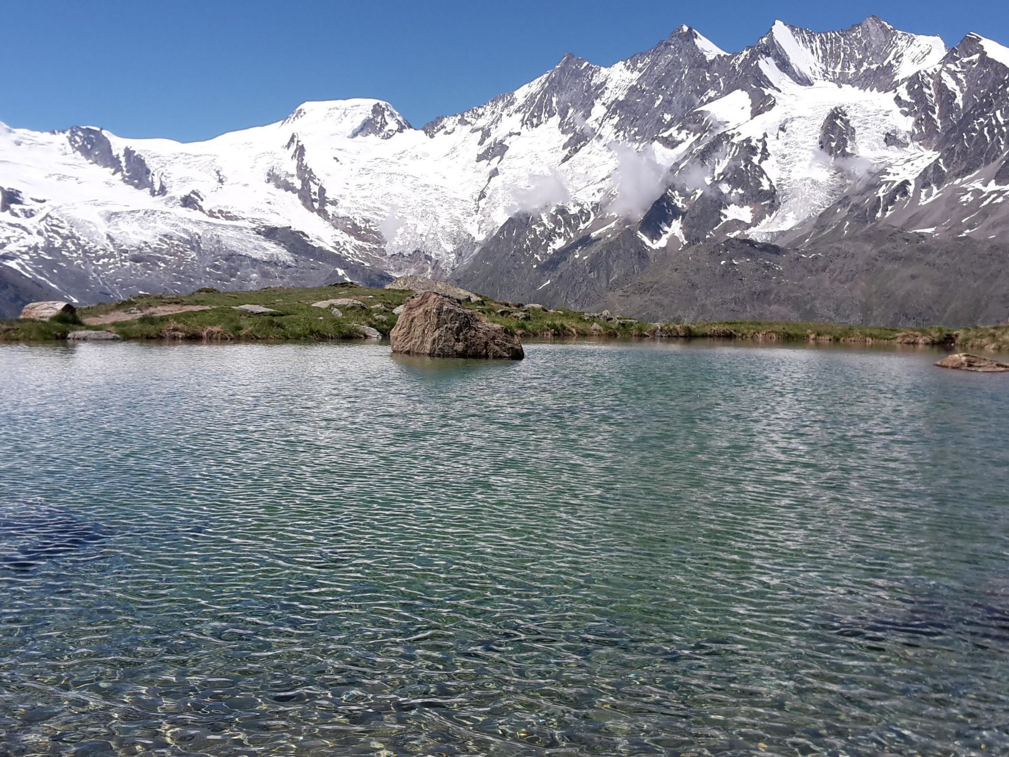 A clear lake with a large rock in the foreground and snow-capped mountains in the background.