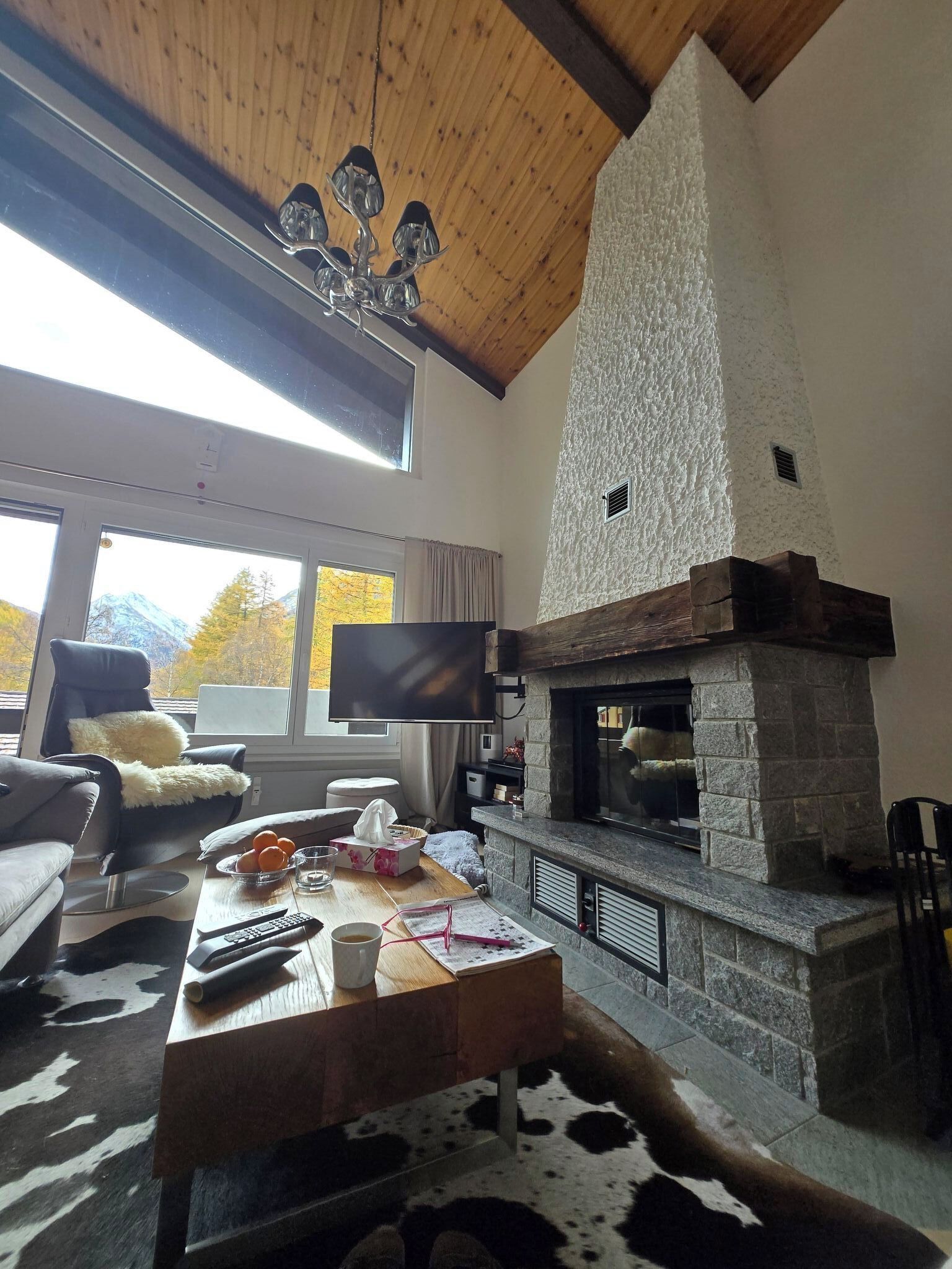A cozy living room with a stone fireplace, a comfortable sofa, a flat-screen TV, and a large window offering a mountain view.