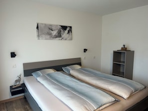 A bedroom with a bed, bedside tables, a nightstand, a wall-mounted lamp, a shelf with decorative items, and a painting. - Ferienhaus Alea Appartement/Fewo 4-Bettwohnung Nr. 20 by Interhome (Saas-Grund)