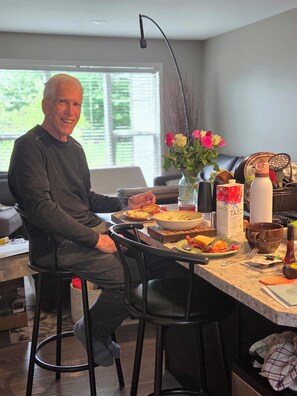 A person seated at a kitchen counter with a dining table, a vase with flowers, and various kitchen items.