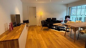 En moderne stue med trægulv, en sort sofa, et lille rundt bord, en lampe og et fjernsyn.