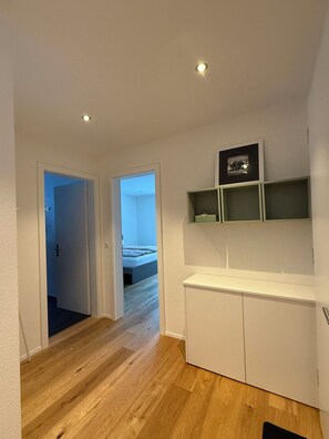 Ein Flur mit Holzboden führt zu einem Schlafzimmer mit Bett und Nachttisch. Über dem Bett befinden sich Einbauschränke und darunter ein weißes Schränkchen.