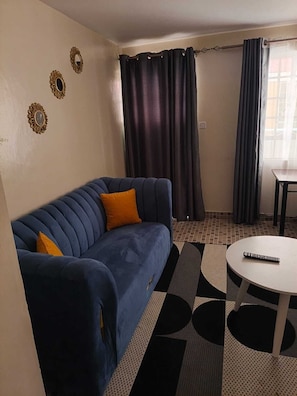 Ein Zimmer mit einem blauen Sofa, einem kleinen runden Tisch, einem gemusterten Teppich und einem Fenster mit Vorhängen.