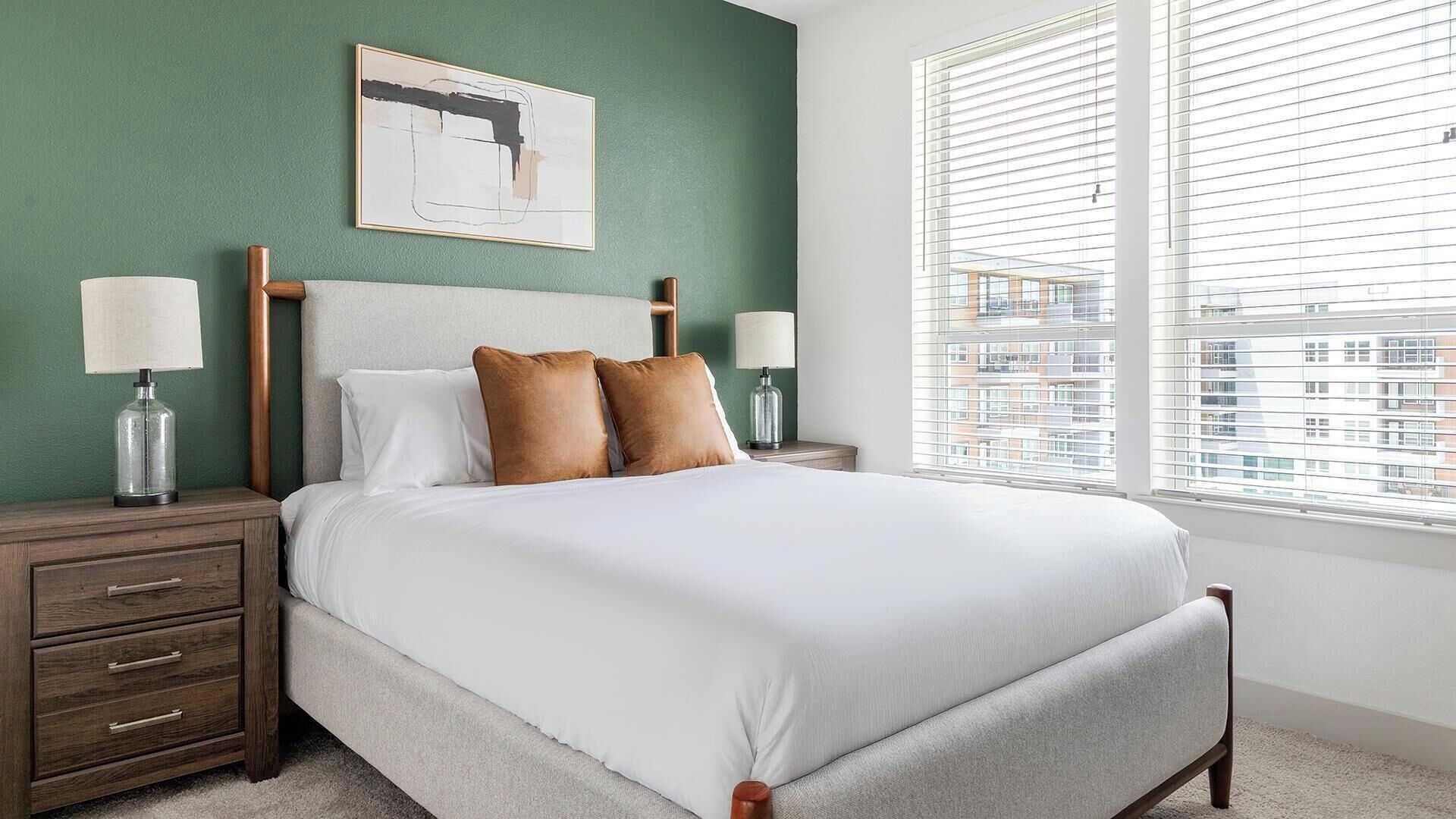 A hotel room with a bed, two bedside tables, a nightstand, a lamp, a framed picture on the wall, and a window with blinds.