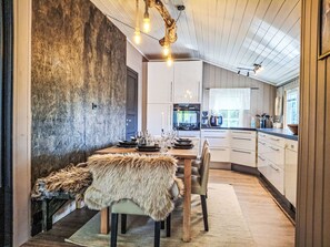 A modern kitchen with white cabinets, a dining area with a fur-covered bench, and a rustic wall. - Hellebu by Interhome (Eikerapen)