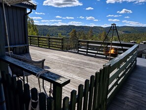 A wooden deck with a wicker chair and a hanging lantern, overlooking a forested mountain range. - Hellebu by Interhome (Eikerapen)