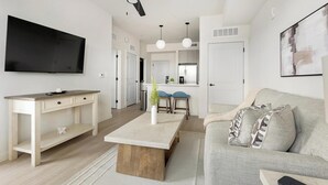 Un salon moderne avec un téléviseur à écran plat, une table basse en bois, un canapé gris et un coin cuisine à l’arrière-plan.