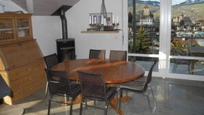 A dining area with a round table and chairs, a wooden cabinet, and a view of a town through large windows. - Ferienwohnung Gassmann Appartement/Fewo 4-Bett Wohnung mit Dusche, Bad u. Balkon by Interhome (Spiez)