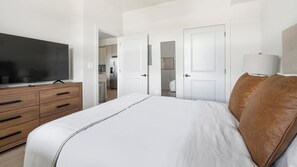 Um quarto com uma cama, uma televisão sobre um aparador de madeira, e uma cozinha visível por uma porta aberta.