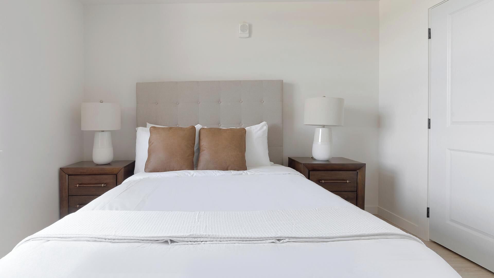 Un lit bien fait, avec une literie blanche, deux tables de chevet avec des lampes et un mur sobre agrémenté d’un interrupteur.