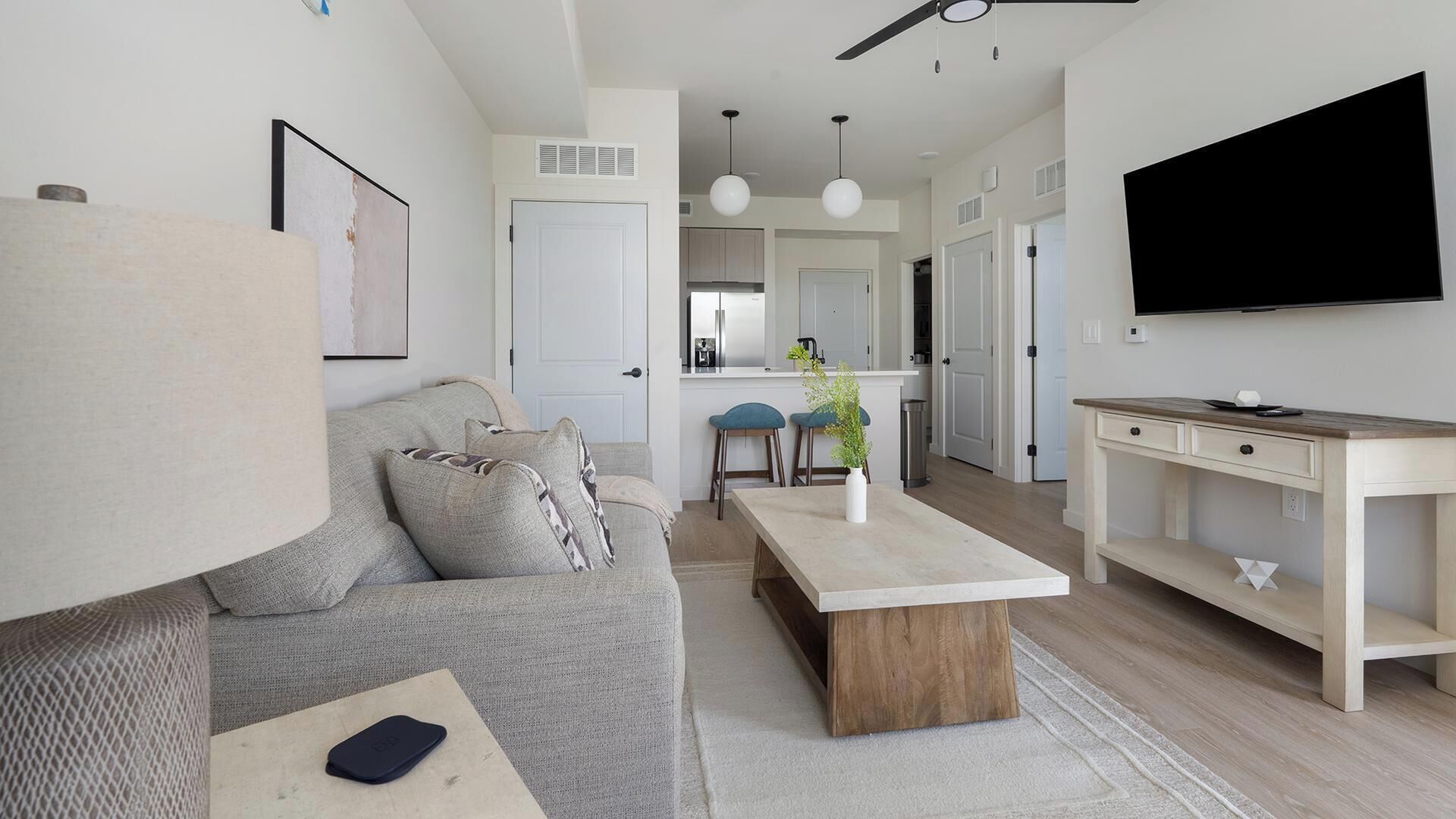 A modern living room with a sofa, coffee table, and TV. There is a kitchen in the background with a fridge and bar stools.