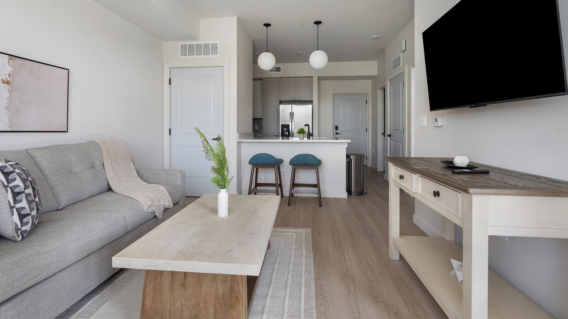 Una sala moderna con un sofá, una mesa de centro y taburetes de bar. Se observa un área de cocina con un refrigerador y un lavamanos.