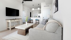 Una sala de estar moderna con un televisor de pantalla plana, una mesa de centro de madera, un sofá gris y una cocina al fondo.
