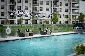 Ein Swimmingpool mit Liegestühlen und Sonnenschirmen, umgeben von einer Glasreling und einem Gebäude im Hintergrund.