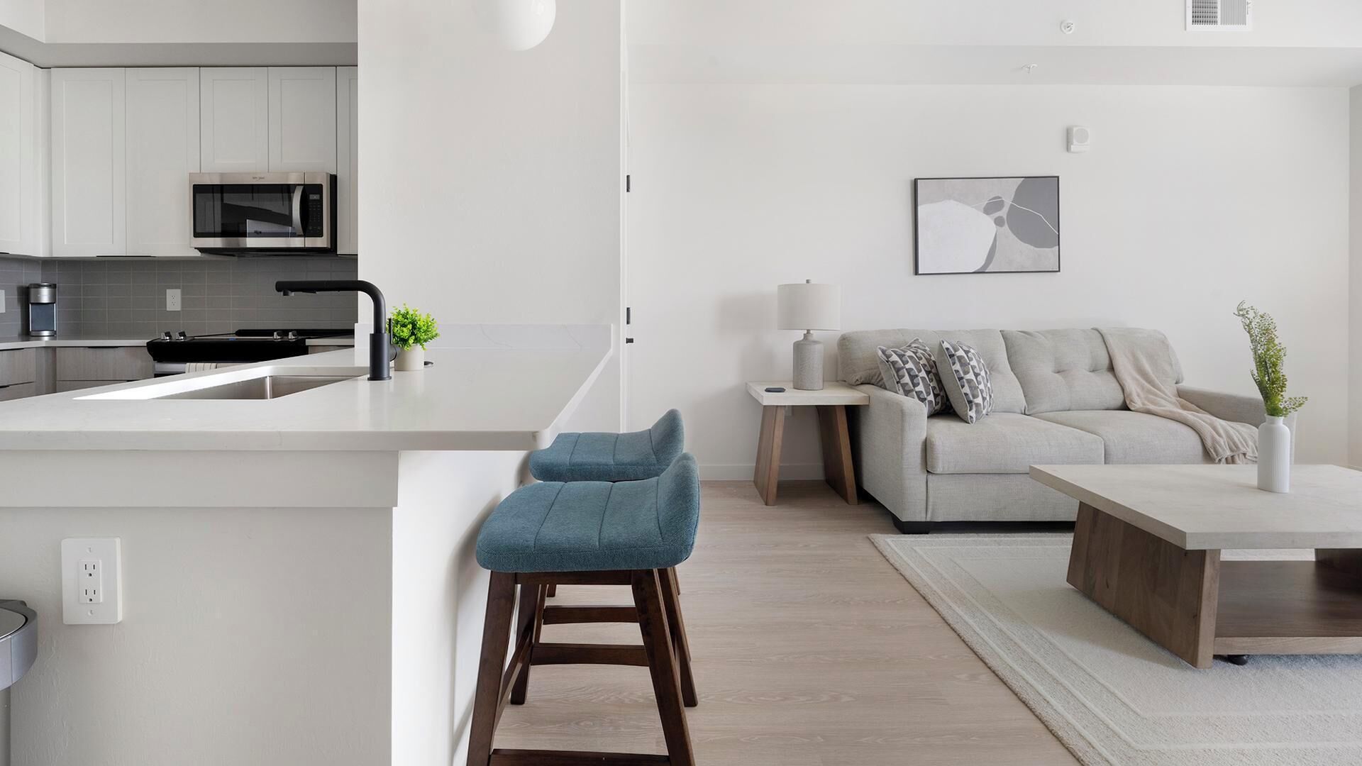 A modern kitchen with a microwave, a sink, and a bar stool. There is a living area with a sofa, a coffee table, and a lamp.