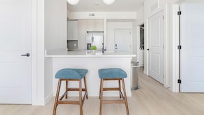 Eine moderne Küche mit weißen Schränken, einem Edelstahl-Kühlschrank und einer weißen Arbeitsplatte. Zwei Holzstühle mit blauen Kissen sind vorhanden.