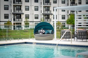 Ein Schwimmbecken mit Glasgeländer, eine Lounge mit einer runden Sitzecke und ein modernes Apartmenthaus im Hintergrund.