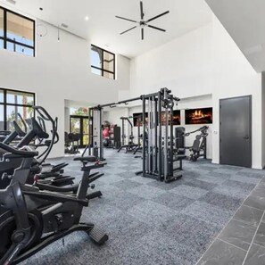 Ein großzügiger Fitnessraum mit unterschiedlichen Trainingsgeräten, großen Fenstern und einem hohen Deckenventilator.