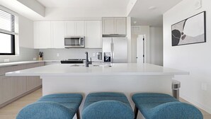 Una cocina moderna con gabinetes blancos, un refrigerador de acero inoxidable, un microondas y una cafetera. Hay tres taburetes tapizados en azul en la isla.