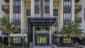 Een modern gebouw met een centrale ingang, meerdere balkons en een parkeerplaats.