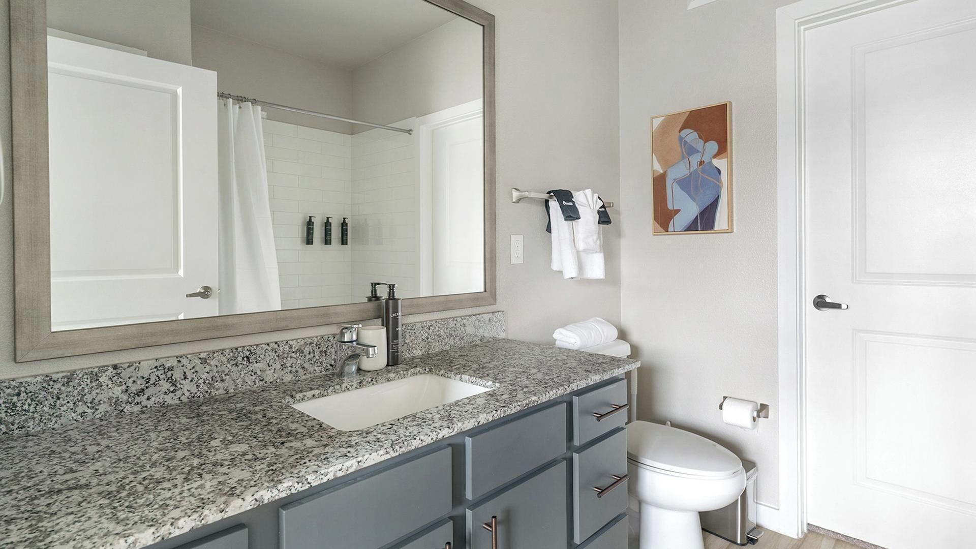 Un baño con encimera de granito, lavamanos blanco, espejo, inodoro y toallero.