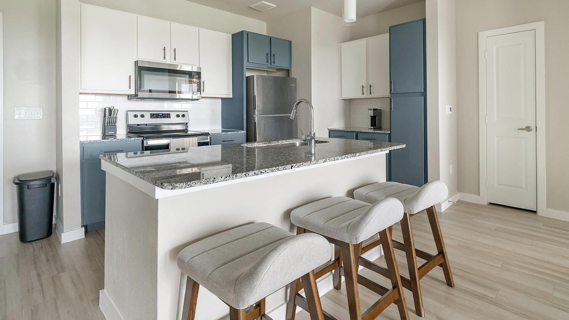 A modern kitchen with white and blue cabinets, stainless steel appliances, a granite countertop, and three bar stools.