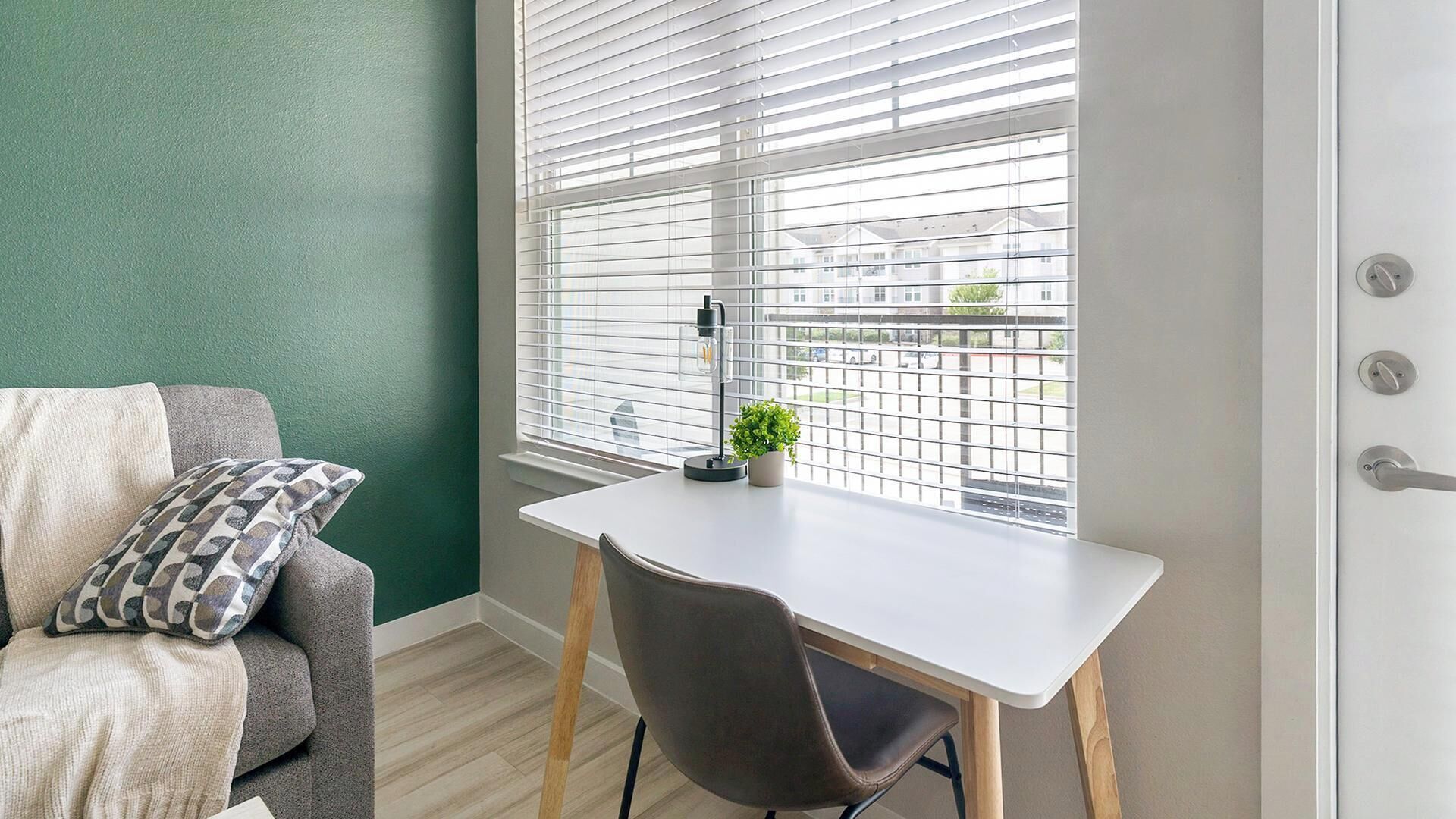 Une pièce avec un bureau blanc, une chaise grise, un mur vert et une fenêtre avec des stores.