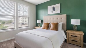 Une chambre d’hôtel avec un grand lit, deux tables de chevet avec des lampes, un mur d’accent vert et une fenêtre avec des stores.