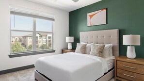 A bedroom with a large bed, two bedside tables with lamps, a window with blinds, and a green accent wall.