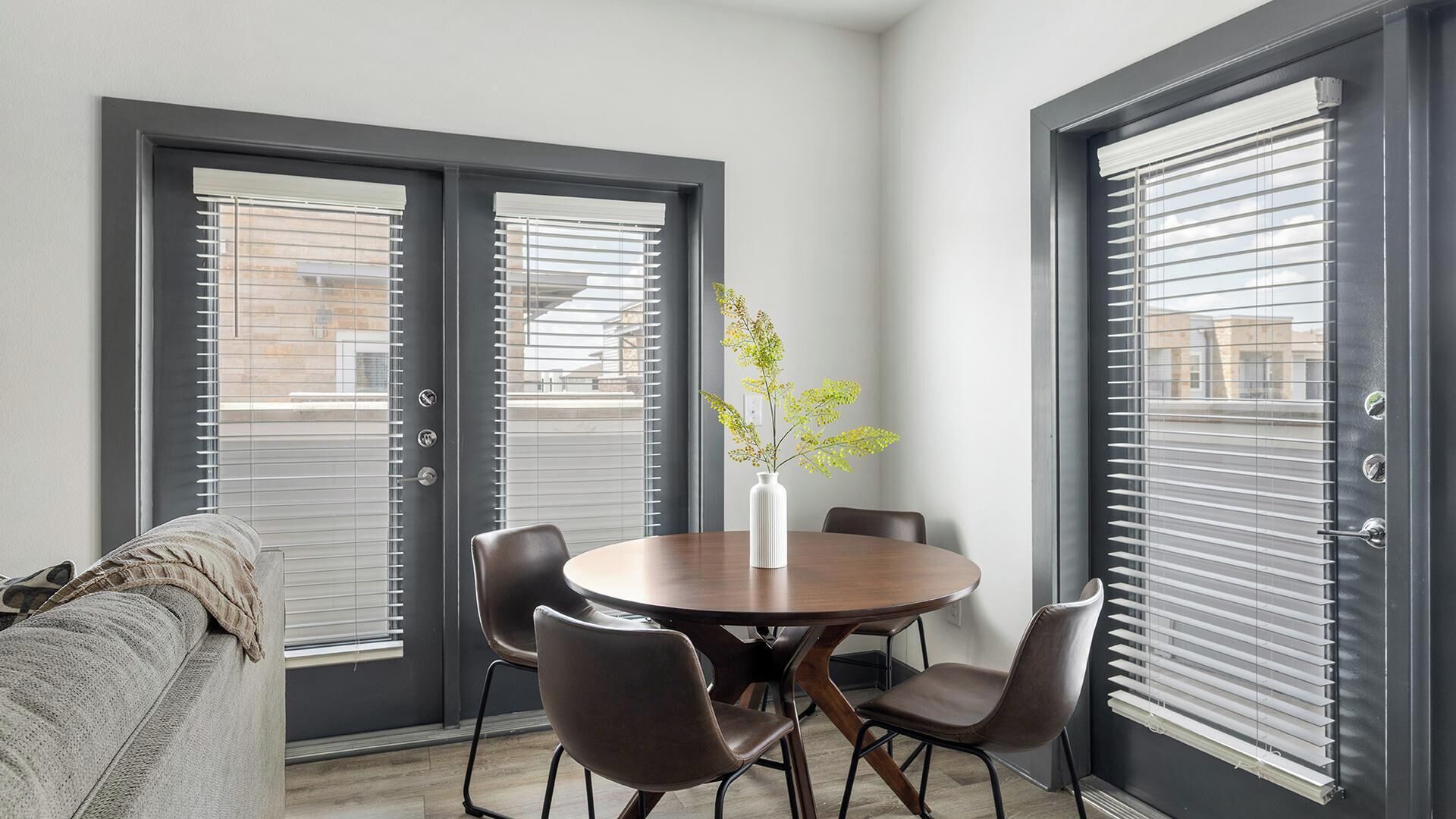 A modern dining area with a round table, four chairs, a vase with flowers, and large windows with blinds.