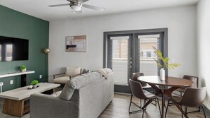 A modern living room with a sofa, a dining table with chairs, a TV mounted on the wall, and a ceiling fan. - Landing | Stunning 3BD, Gym (Terrell)