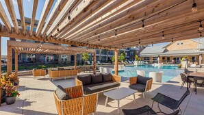 Un espacio patio al aire libre con piscina, pérgola de madera, mobiliario de mimbre y vista del edificio al fondo.