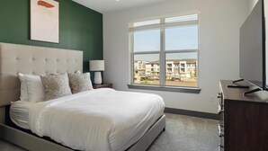 Un dormitorio con cama, mesita de noche, lámpara, televisor y ventana con vistas a edificios.
