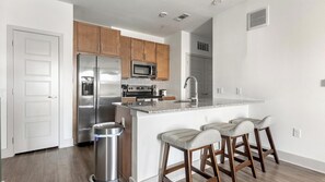 A modern kitchen with stainless steel appliances, wooden cabinets, and a bar stool seating area. - Landing | Amazing 1BD, Clubhouse, Gym (Leander)