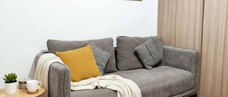 Un sofá gris con cojines estampados, una mesita con una planta y una taza, y una pared decorada con ilustraciones de estilo vintage.