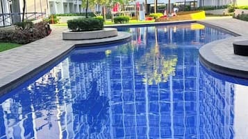 A swimming pool with a tiled edge, surrounded by a landscaped area with trees and a building in the background.