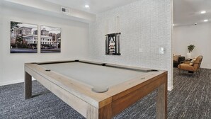 Ein Billardtisch in einem Raum mit strukturierter Wand, zwei gerahmte Bilder und eine Sitzecke mit Sessel und Sofa.