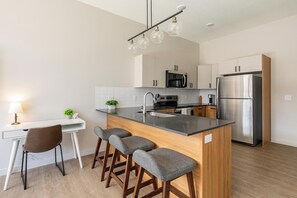 A modern kitchen with a central island, bar stools, a dining area with a chair, and a refrigerator. - Landing | Modern 1BD, Game Room, Gym (Salt Lake City)