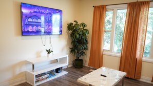 Un salon avec un téléviseur fixé au mur, un meuble de rangement blanc, une table basse en marbre, une plante en pot et une fenêtre avec des rideaux.