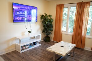 A living room with a TV mounted on the wall, a white entertainment unit, a marble coffee table, a potted plant, and a window with curtains. - Peaceful Kitchener Getaway 5BR Home w/ BBQ & Modern Kitchen (Kitchener)