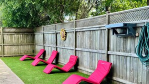 Un jardin avec une clôture en bois, un chemin en béton et quatre chaises longues roses.