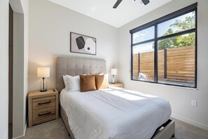 Ein Schlafzimmer mit einem großen Bett, zwei Nachttischen, einem Deckenventilator, einem Fenster mit Blick auf einen Holenzaun und einem gerahmten Bild an der Wand.