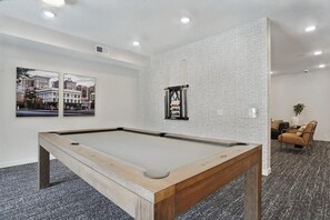Ein Billardtisch in einem Raum mit strukturierter Wand, zwei gerahmte Bilder und eine Sitzecke mit Sessel und Sofa.