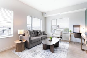 Ein modernes Wohnzimmer mit einem grauen Sofa, einem runden Couchtisch, einem weißen Sessel und einem Schreibtisch mit einem Computer. Zwei Fenster mit Jalousien, eine Lampe auf einem Beistelltisch und eine kleine Pflanze auf dem Couchtisch sind ebenfalls vorhanden.