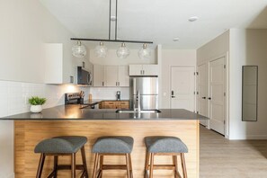 A modern kitchen with a central island, bar stools, a refrigerator, and a pendant light fixture. - Landing | Modern 1BD, Yoga Studio, Game Room (Salt Lake City)