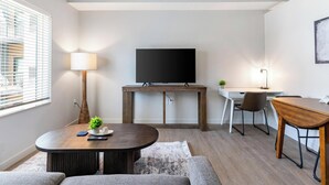 Una sala de estar moderna con un televisor de pantalla plana, una mesa de centro de madera, un sofá, un escritorio con una silla y una lámpara.
