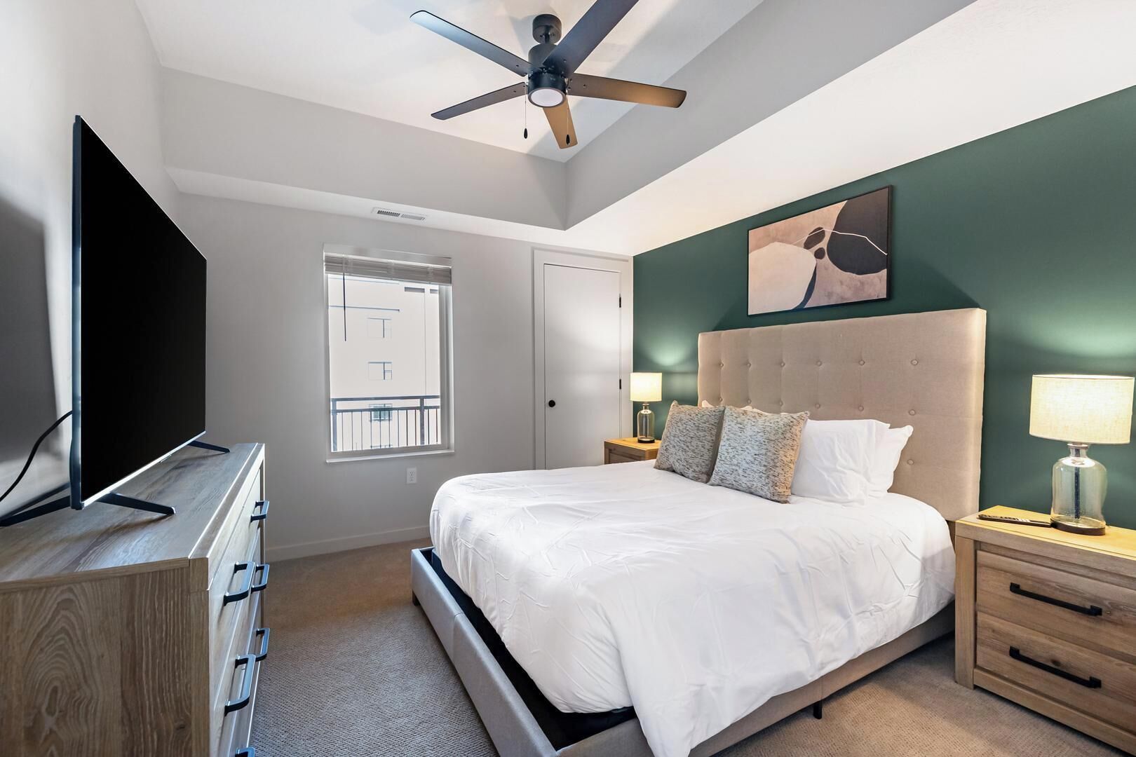 Een moderne slaapkamer met een bed, een televisie, een plafondventilator, een raam en een nachtkastje met een lamp.